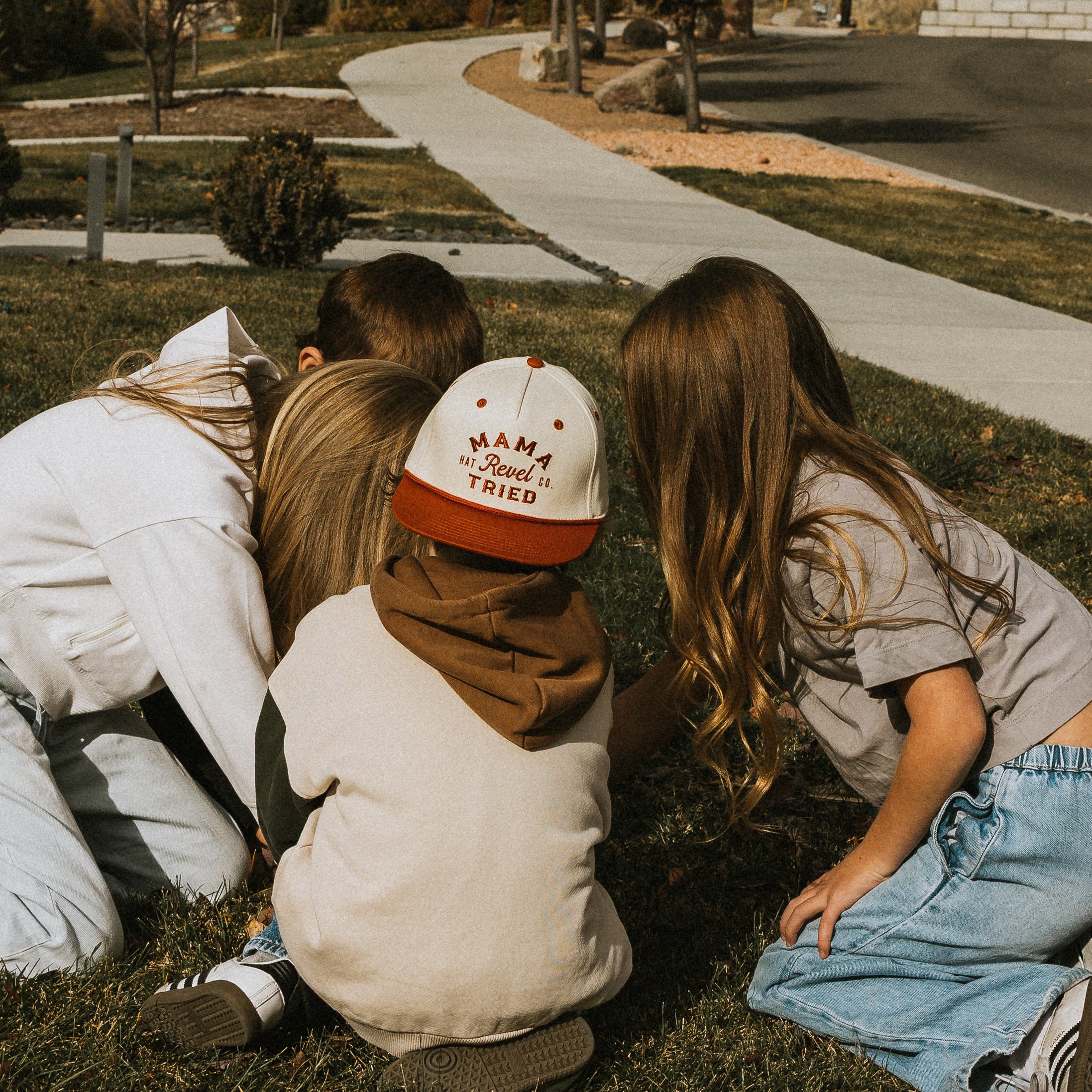 Children playing on a grassy area one wearing  a snapback hat that says "mama tried" on it. 