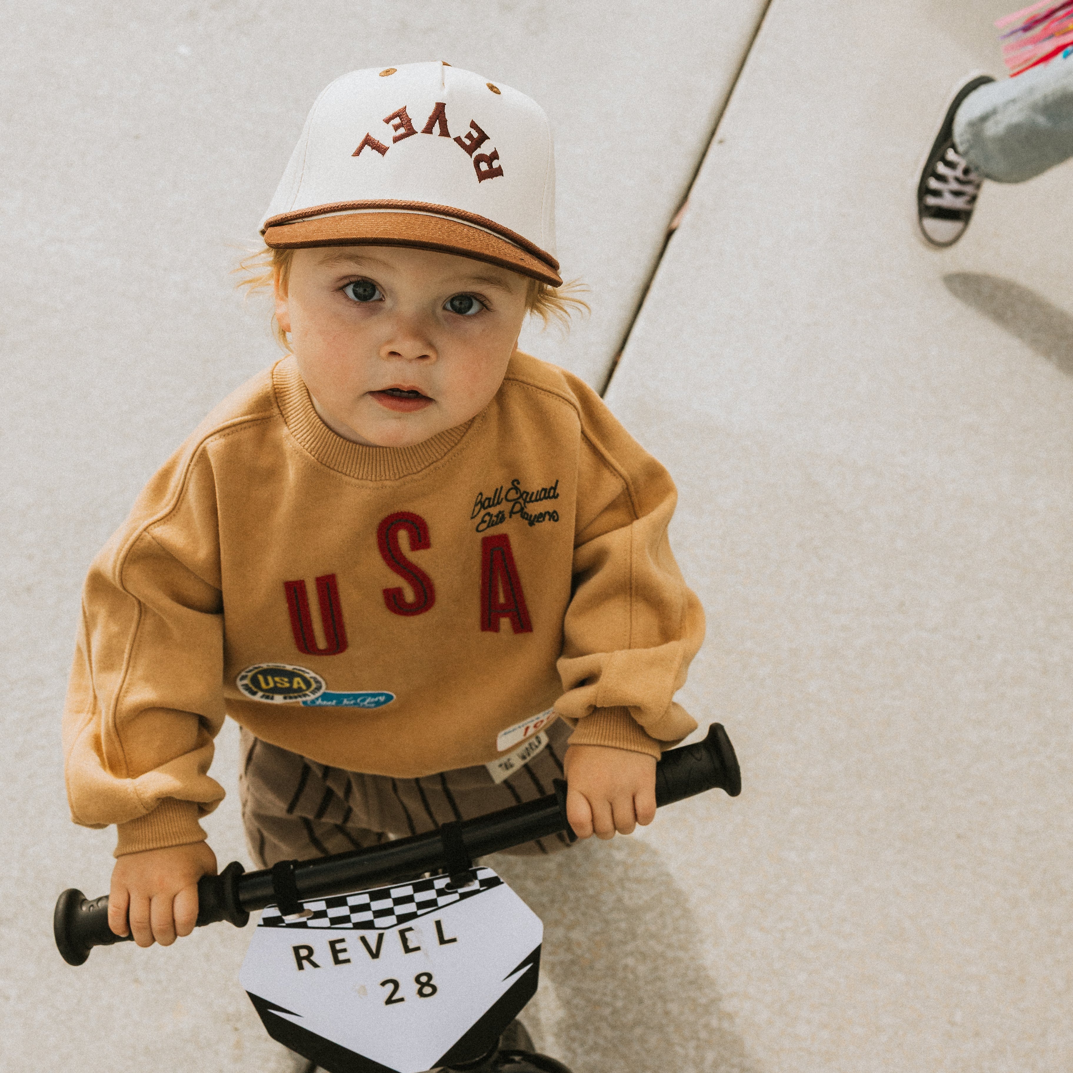 Child wearing a snapback cap and sweatshirt, holding a bike with a checkered flag design.
