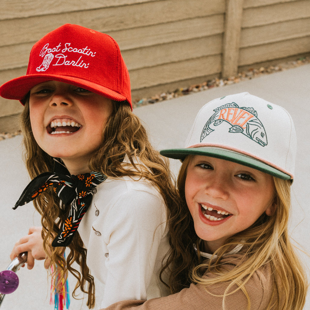 Two children wearing baseball caps, one red and one with a fish graphic, sitting outdoors.