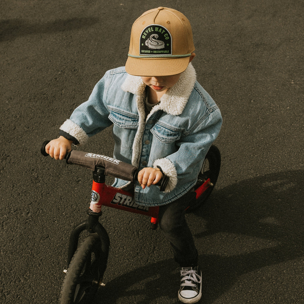 Child wearing a tan cap with a snake graphic, riding a Strider balance bike.