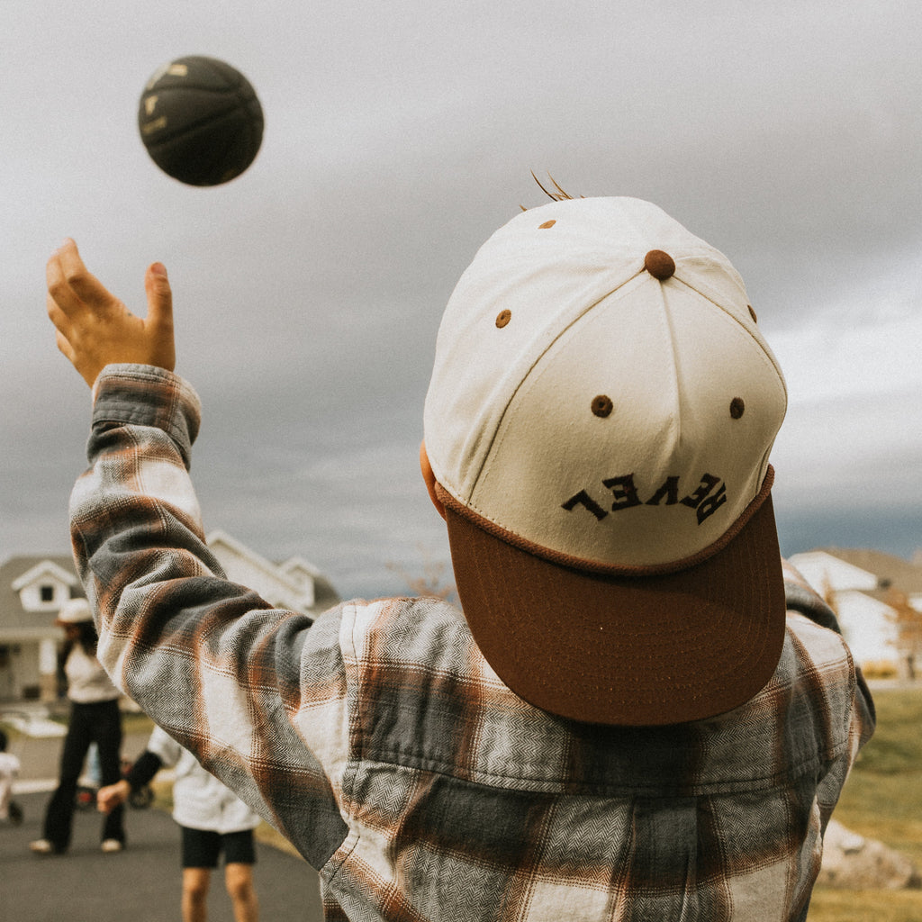 Kids wearing a snapback cap and plaid shirt, reaching out to catch a basketball on a cloudy day.