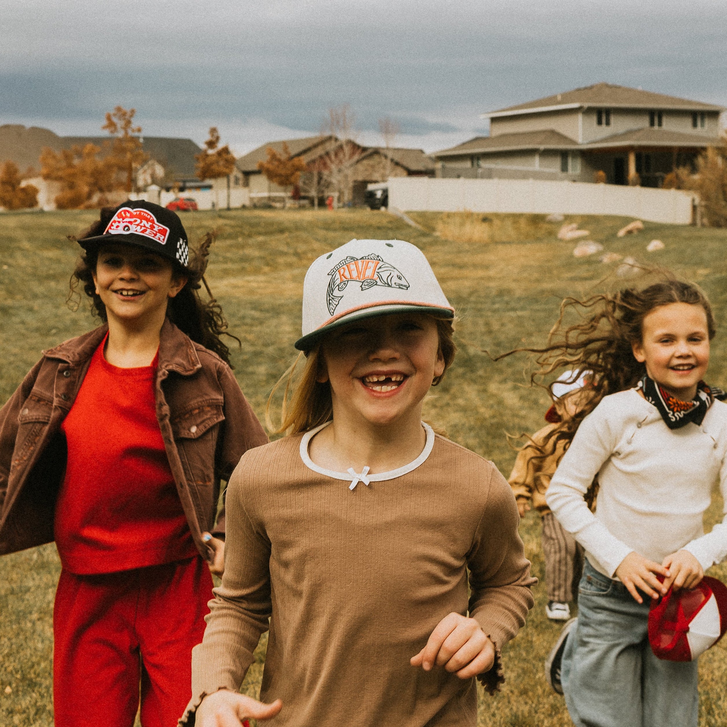 Three children running in a grassy field on a cloudy day wearing snapback hats