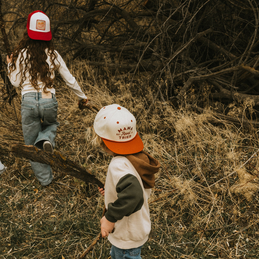 Two children in a forest setting, wearing snapback caps and playing with sticks.