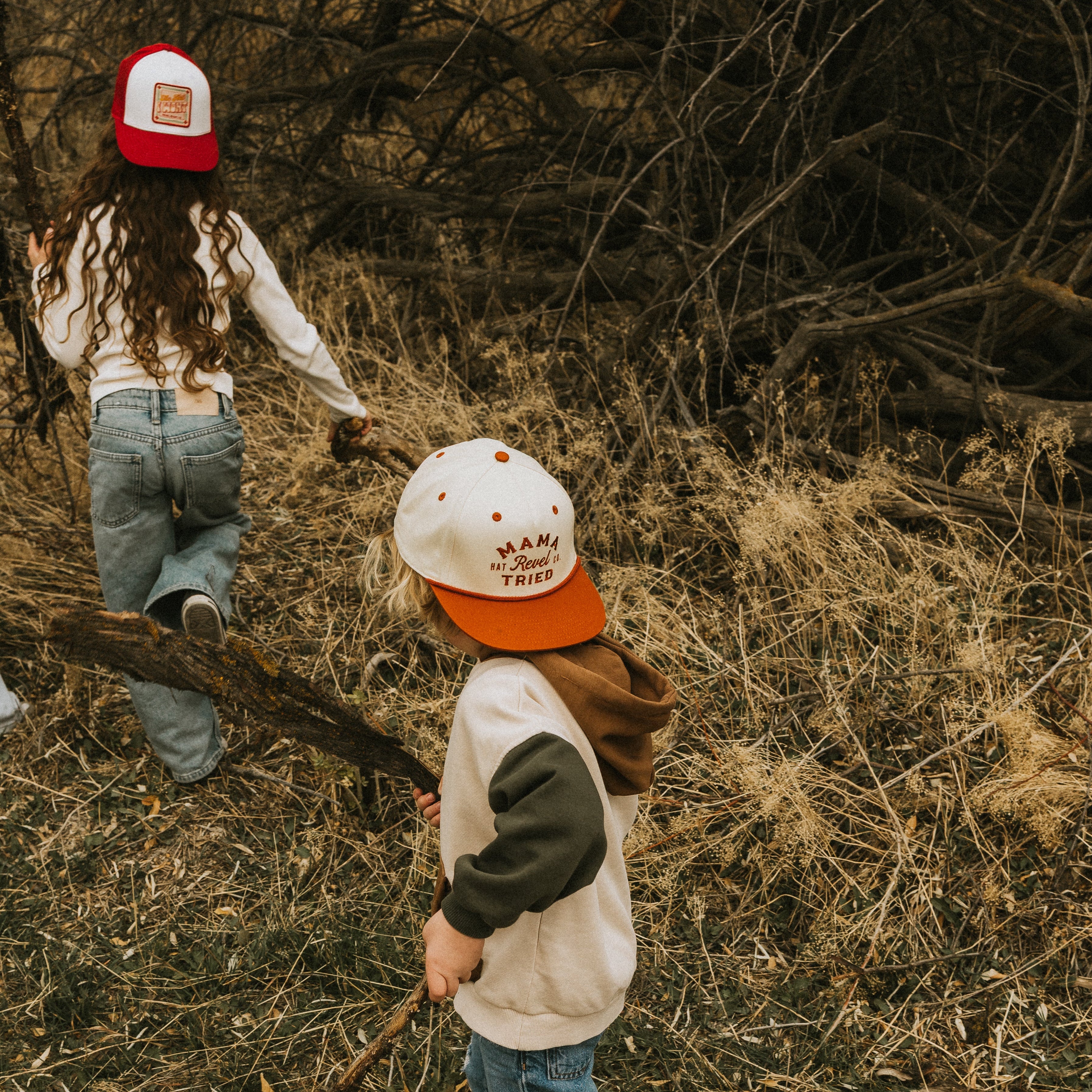 Two children in a forest setting, wearing snapback caps and playing with sticks.