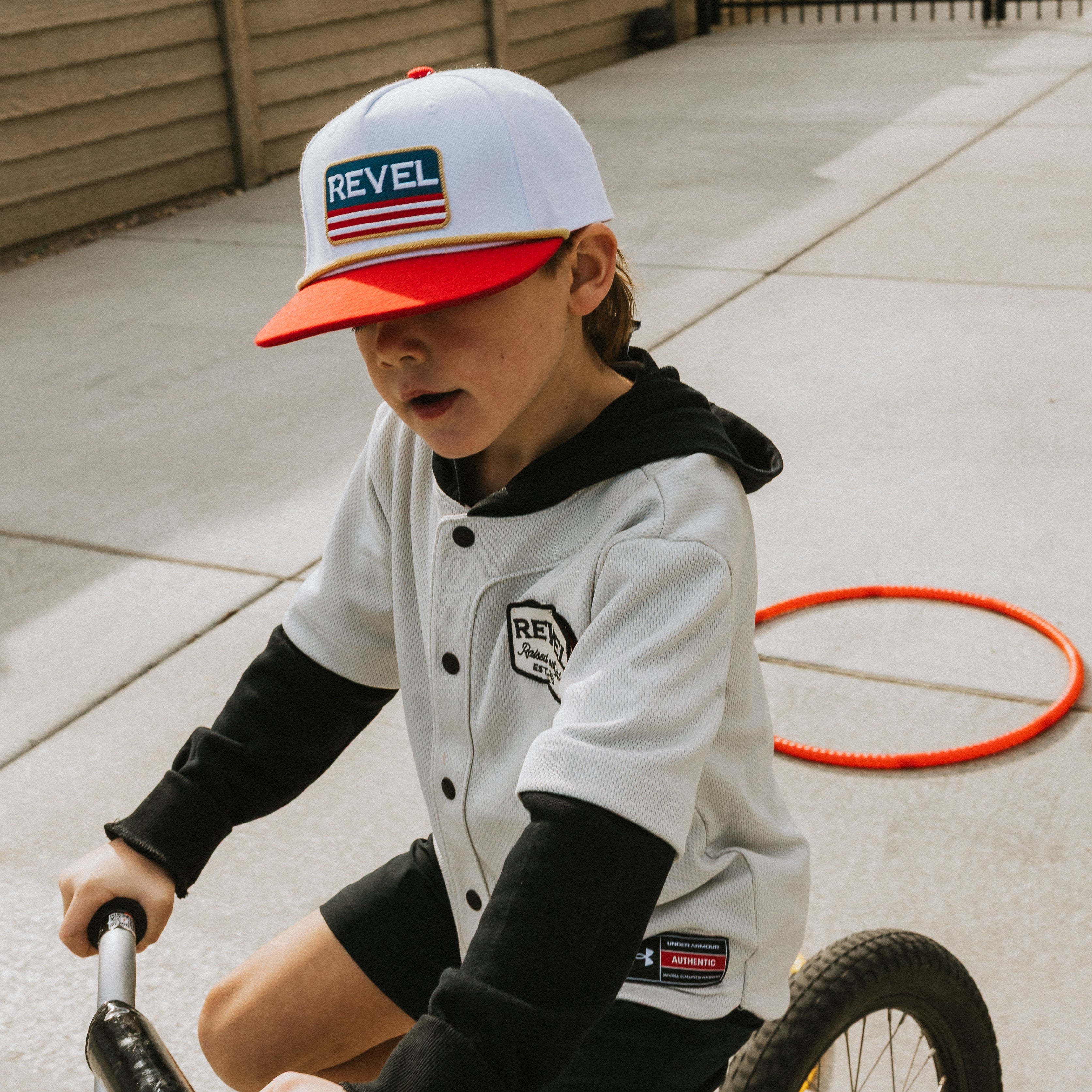 Child riding a tricycle wearing a Red American Flag baseball cap and jacket in an outdoor setting.