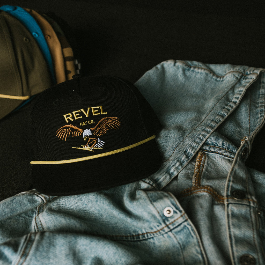 Black cap with an embroidered 'REVEL' logo and eagle on a denim jacket against a dark background