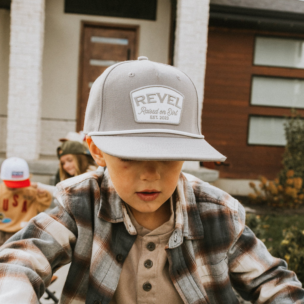 Child wearing a grey 'Revel' snapback cap outdoors with a building in the background