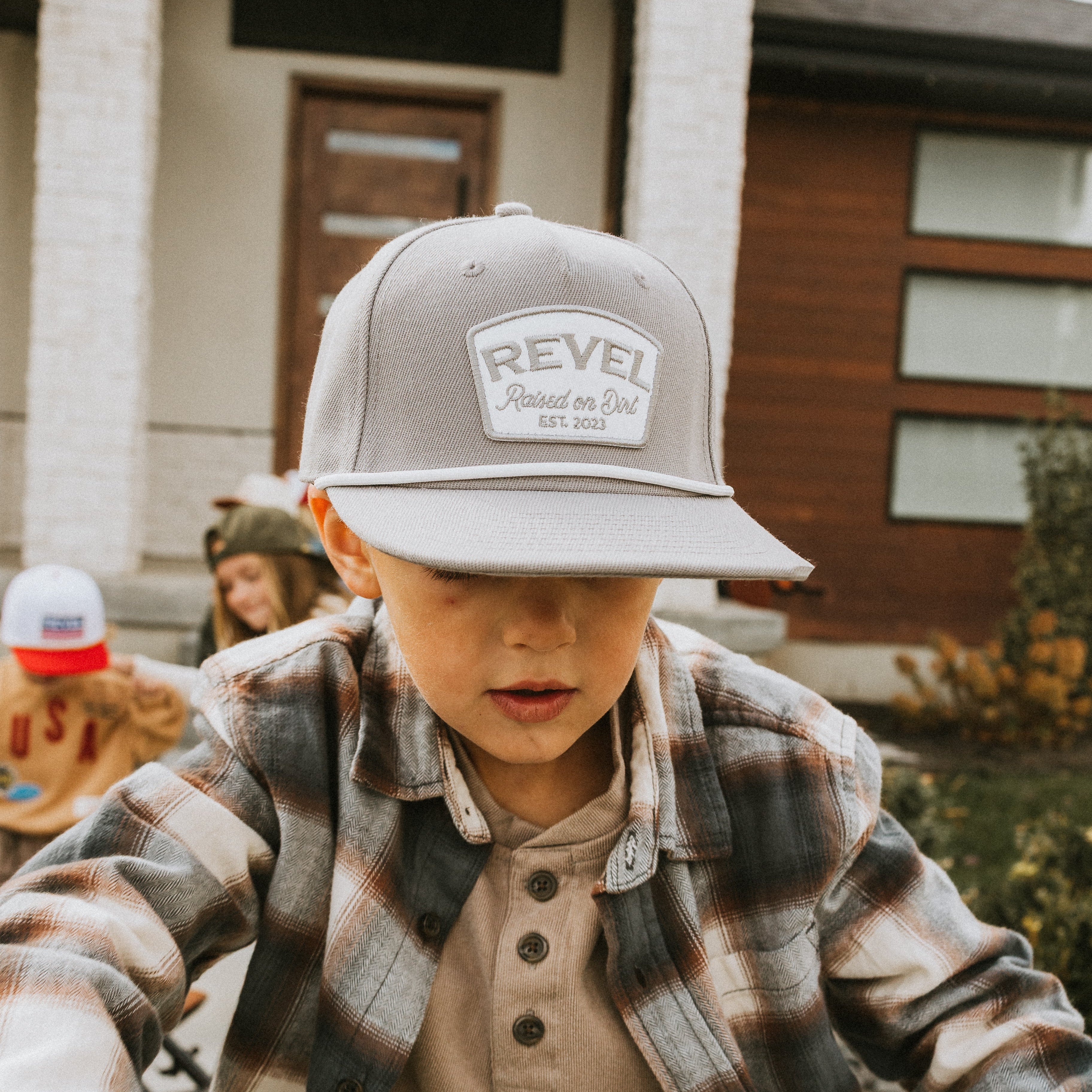 Child wearing a grey 'Revel' snapback cap outdoors with a building in the background