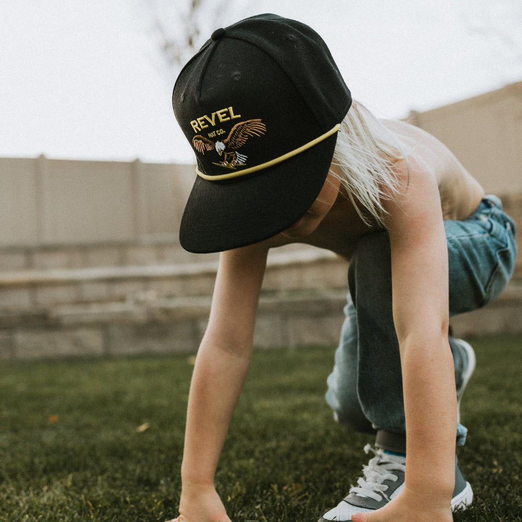 Kid playing in grass wearing a vintage inspired snapback that is black with an embroidered eagle and yellow rope and embroidered logo. 