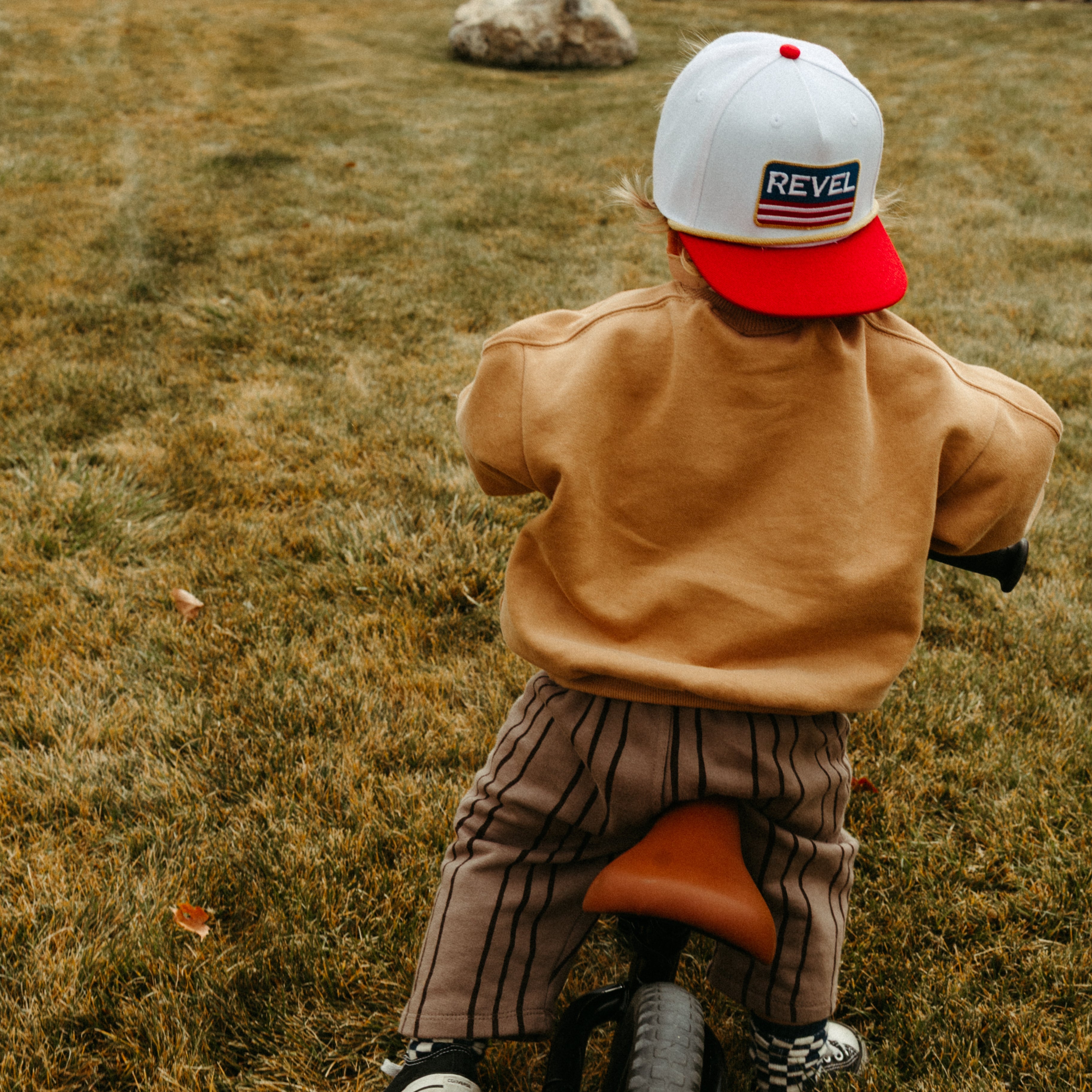 Child wearing a red and white snapback hat riding a bike in a grassy field.